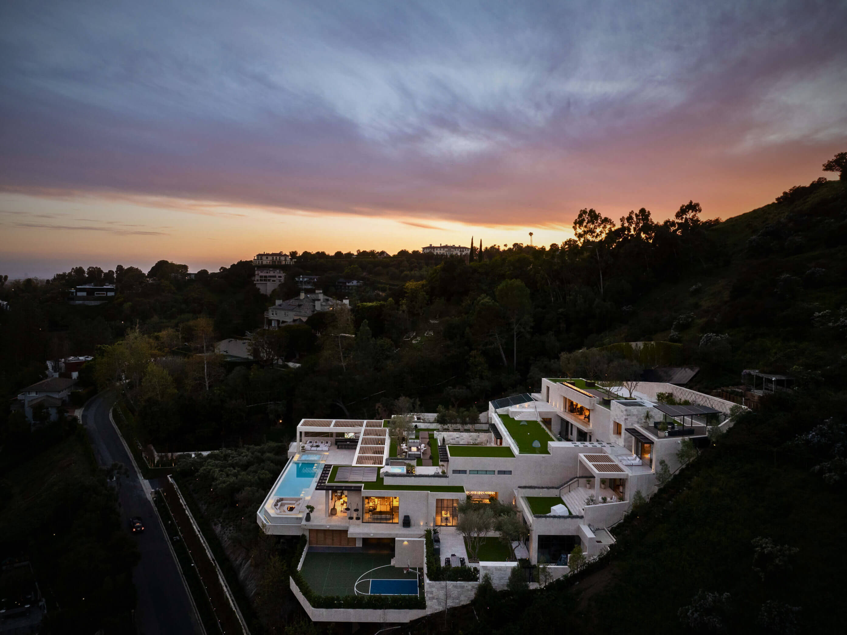 Aerial view of a modern hillside mansion at sunset with green rooftops, solar panels and outdoor living spaces